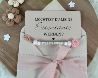 May include: A pink envelope with a white card that says "Möchtest du meine Patentante werden?"  The card is tied with a pink ribbon and a beaded keychain with the word "Patentante" spelled out in white letters.