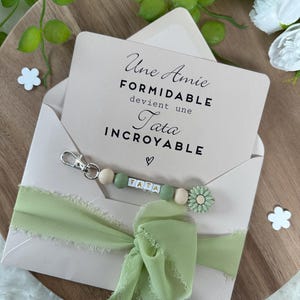 Puede incluir: Un sobre rosa claro con una cinta verde y un llavero con cuentas de madera y un dije de flor. El sobre contiene una tarjeta con el texto "Une Amie Formidable devient une Tata Incroyable".