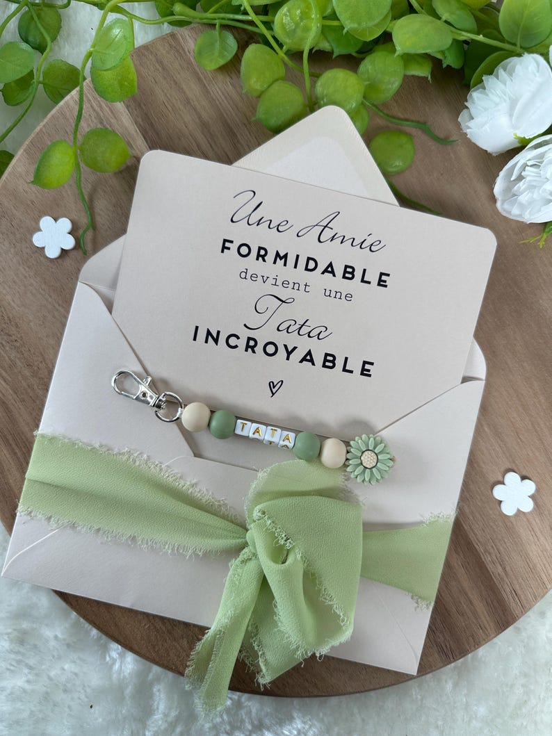 Puede incluir: Un sobre rosa claro con una cinta verde y un llavero con cuentas de madera y un dije de flor. El sobre contiene una tarjeta con el texto "Une Amie Formidable devient une Tata Incroyable".