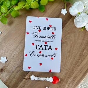 以下が含まれることがあります： 赤いハートのアクセントとフランス語のテキストが特徴の白いカード。「UNE SOEUR Formidable devient toujours une TATA Exceptionnelle」と書かれています。「TATA」という単語が入ったビーズのキーホルダーも表示されています。木製のトレイにアイテムが置かれています。