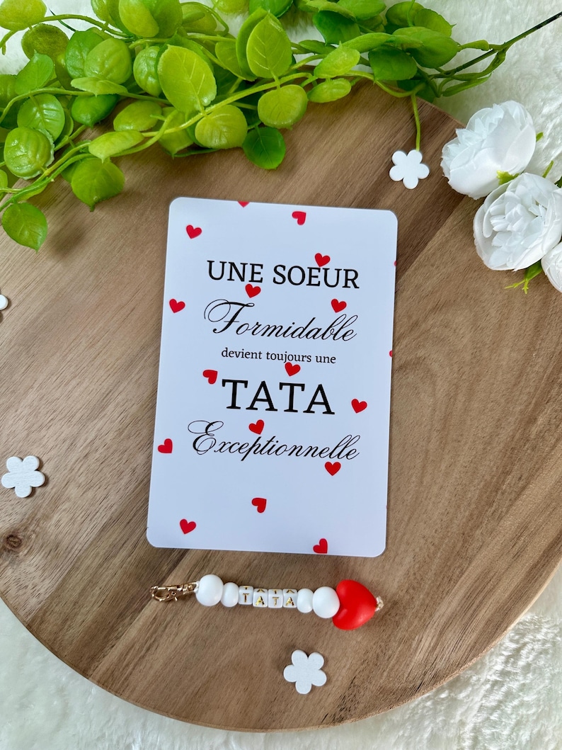 以下が含まれることがあります： 赤いハートのアクセントとフランス語のテキストが特徴の白いカード。「UNE SOEUR Formidable devient toujours une TATA Exceptionnelle」と書かれています。「TATA」という単語が入ったビーズのキーホルダーも表示されています。木製のトレイにアイテムが置かれています。
