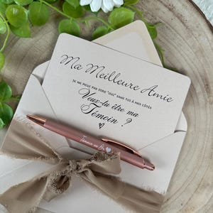 K&ouml;nnte beinhalten: Eine hellrosa Einladungskarte mit dem Text "Ma Meilleure Amie" und einem passenden Stift. Die Karte befindet sich in einem hellrosa Umschlag, der mit einem beigen Band gebunden ist. Eine wei&szlig;e G&auml;nsebl&uuml;mchen und Gr&uuml;nzeug sind im Hintergrund.