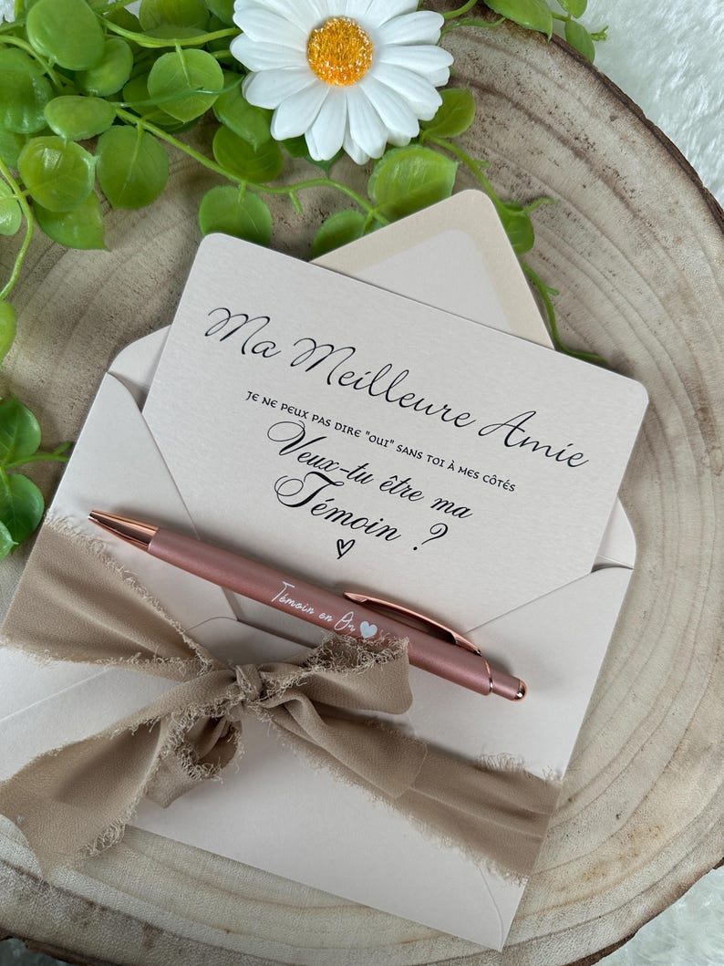 K&ouml;nnte beinhalten: Eine hellrosa Einladungskarte mit dem Text "Ma Meilleure Amie" und einem passenden Stift. Die Karte befindet sich in einem hellrosa Umschlag, der mit einem beigen Band gebunden ist. Eine wei&szlig;e G&auml;nsebl&uuml;mchen und Gr&uuml;nzeug sind im Hintergrund.