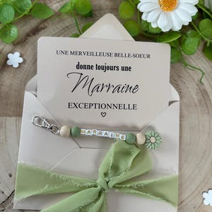 May include: A light brown card with the text "Une merveilleuse belle-soeur donne toujours une Marraine exceptionnelle" and a green and white beaded keychain with the word "Marraine" spelled out on it. The keychain is attached to a green ribbon bow.