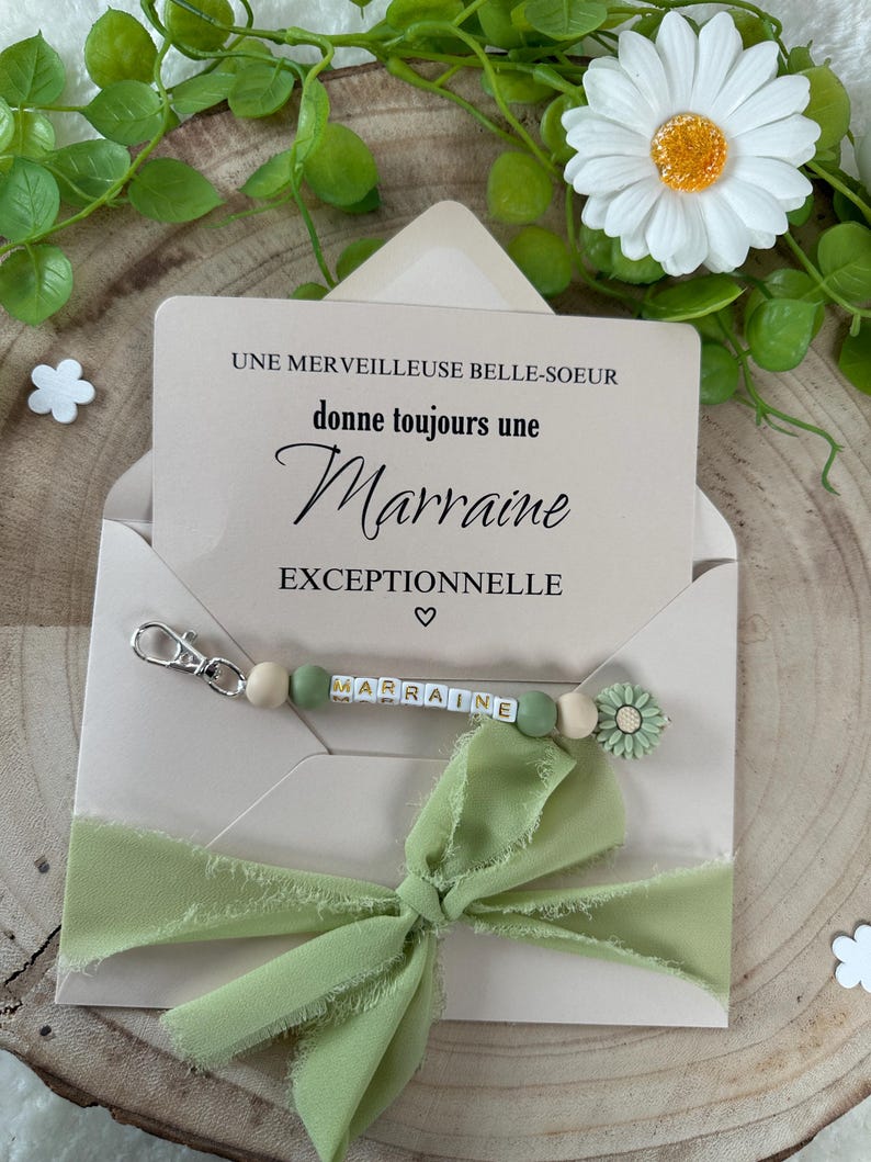 May include: A light brown card with the text "Une merveilleuse belle-soeur donne toujours une Marraine exceptionnelle" and a green and white beaded keychain with the word "Marraine" spelled out on it. The keychain is attached to a green ribbon bow.