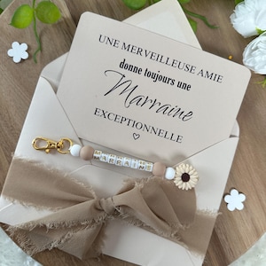 以下が含まれることがあります： 「UNE MER VEILLEUSE AMIE donne toujours une Marraine EXCEPTIONNELLE」というテキストとハートマークが書かれた白いカード。ゴールドの文字で「MARRAINE」と書かれたキーホルダーと白いひまわりのチャームが付いた茶色のリボン。
