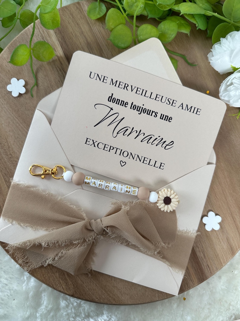 以下が含まれることがあります： 「UNE MER VEILLEUSE AMIE donne toujours une Marraine EXCEPTIONNELLE」というテキストとハートマークが書かれた白いカード。ゴールドの文字で「MARRAINE」と書かれたキーホルダーと白いひまわりのチャームが付いた茶色のリボン。