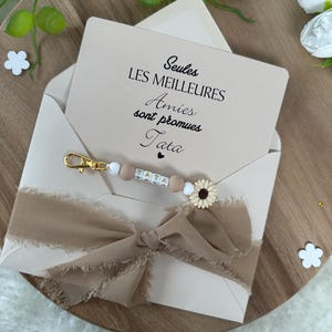 May include: A white card with the text "Seules les meilleures Amies sont promues Tata" in black lettering. A brown ribbon is tied around the card, and a beaded keychain with a sunflower charm is on top of the ribbon.
