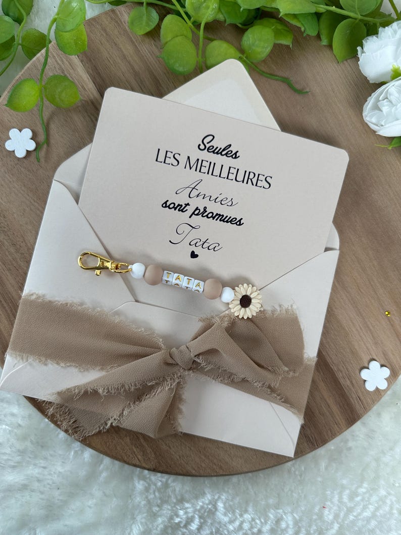 May include: A white card with the text "Seules les meilleures Amies sont promues Tata" in black lettering. A brown ribbon is tied around the card, and a beaded keychain with a sunflower charm is on top of the ribbon.