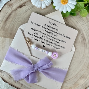 May include: A light beige envelope with a lavender ribbon and a beaded pacifier clip. The clip has the name "TATA" spelled out. A card inside the envelope reads "Breaking news: You're getting a family promotion..." and is decorated with a daisy.