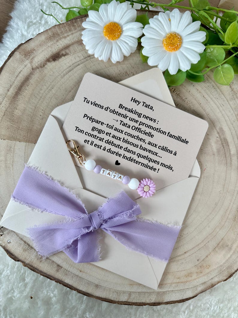 May include: A light beige envelope with a lavender ribbon and a beaded pacifier clip. The clip has the name "TATA" spelled out. A card inside the envelope reads "Breaking news: You're getting a family promotion..." and is decorated with a daisy.