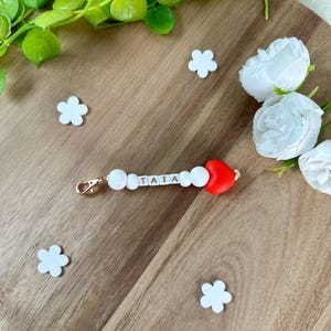 以下が含まれることがあります： 「TATA」と書かれた白いビーズと赤いハートのチャームが付いたキーホルダー。キーホルダーには金色の留め金が付いています。背景には白い花の形の装飾と白いバラが木製の表面にあります。