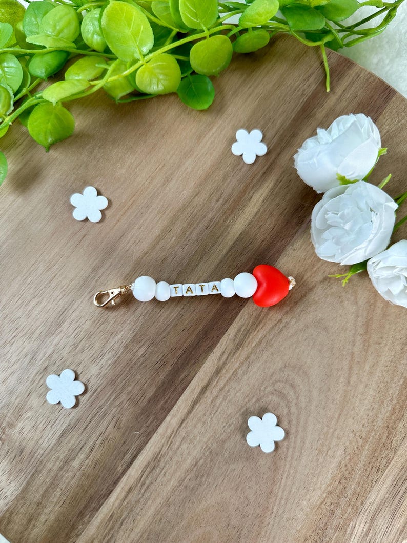 以下が含まれることがあります： 「TATA」と書かれた白いビーズと赤いハートのチャームが付いたキーホルダー。キーホルダーには金色の留め金が付いています。背景には白い花の形の装飾と白いバラが木製の表面にあります。