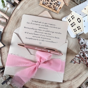 Puede incluir: Un sobre beige claro con una cinta rosa y un bolígrafo de oro rosa. El sobre contiene una tarjeta con texto en francés. El bolígrafo tiene la inscripción "Marraine d'amour". Hay casas y estrellas de madera decorativas en el fondo.