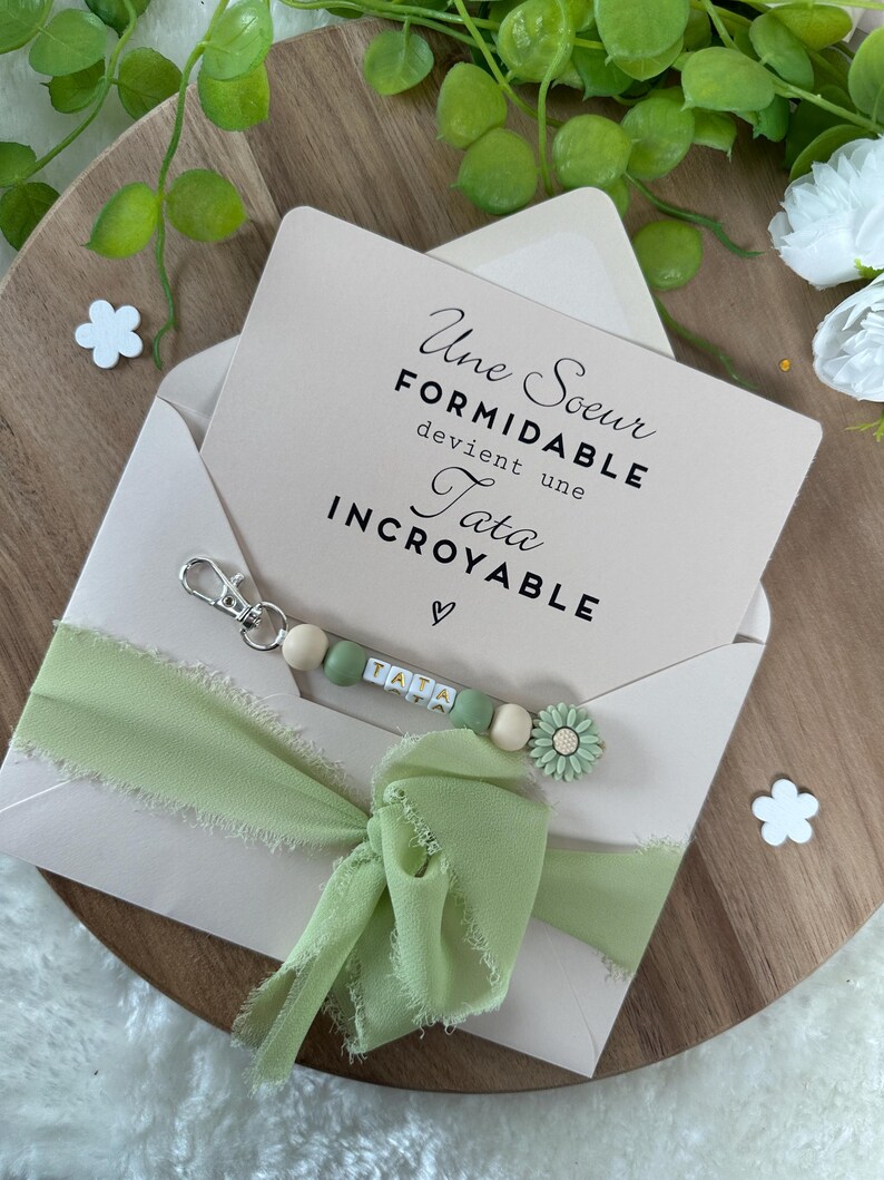 Puede incluir: Un sobre verde claro con una tarjeta en su interior que dice "Une Soeur Formidable devient une Tata Incroyable". Una cinta verde claro est&aacute; atada alrededor del sobre y un llavero de cuentas verde claro y blanco con la palabra "Tata" deletreada est&aacute; unido a la cinta.