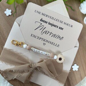 以下が含まれることがあります： 白いカードに「Une merveilleuse soeur donne toujours une Marraine exceptionnelle」というテキストとハートのシンボルが書かれています。茶色のリボンがカードの周りに結ばれています。白いビーズと茶色のビーズでできたキーホルダーに、金色の文字で「Marraine」と書いてあります。キーホルダーはカードの上に置かれています。