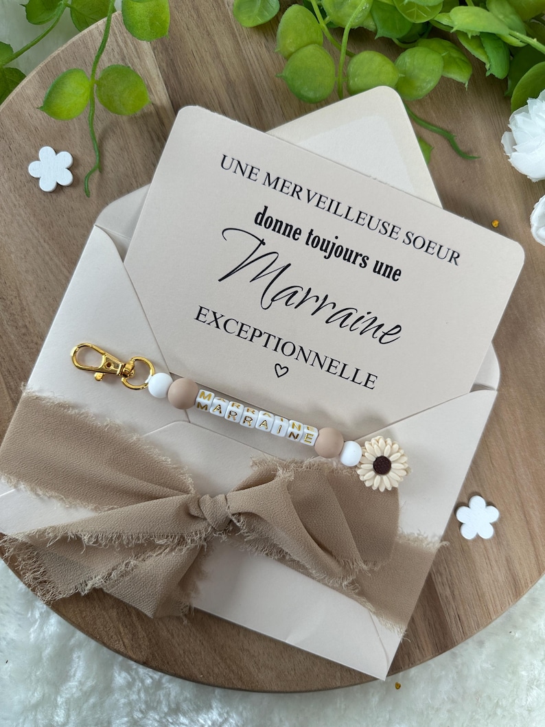 以下が含まれることがあります： 白いカードに「Une merveilleuse soeur donne toujours une Marraine exceptionnelle」というテキストとハートのシンボルが書かれています。茶色のリボンがカードの周りに結ばれています。白いビーズと茶色のビーズでできたキーホルダーに、金色の文字で「Marraine」と書いてあります。キーホルダーはカードの上に置かれています。