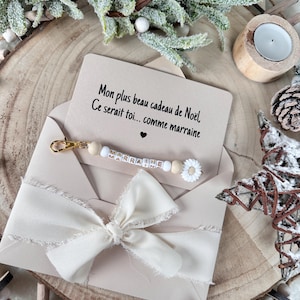Puede incluir: Un sobre beige con una cinta crema y una tarjeta con texto en francés. Un llavero con la palabra "MARRAINE" está adjunto. La escena está decorada con elementos festivos.