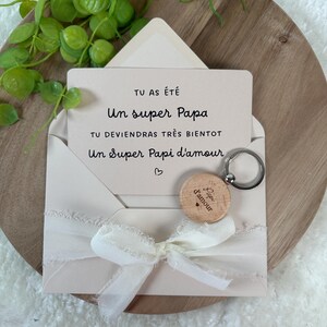 May include: A light pink envelope with a white ribbon bow, holding a card with French text and a wooden keychain. The keychain has the words "Papi d'amour" engraved on it. The card reads "Un super Papa".