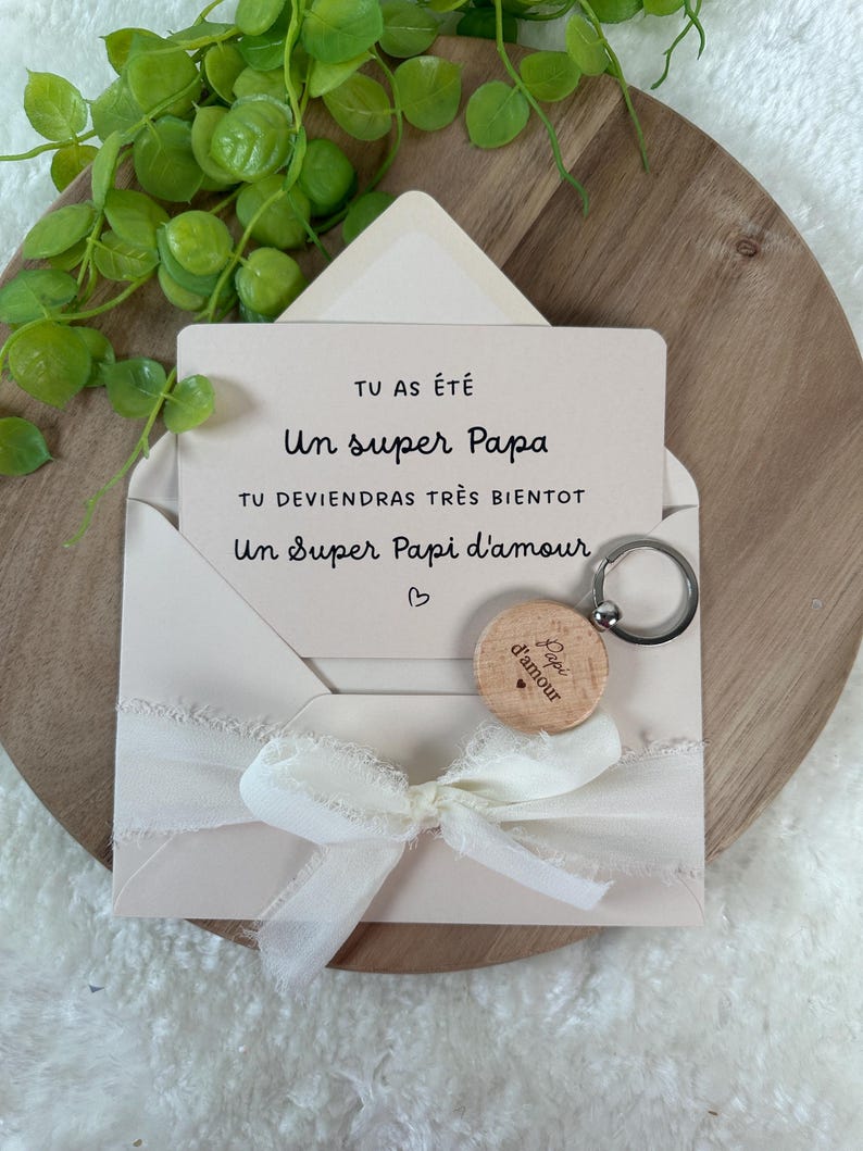 May include: A light pink envelope with a white ribbon bow, holding a card with French text and a wooden keychain. The keychain has the words "Papi d'amour" engraved on it. The card reads "Un super Papa".