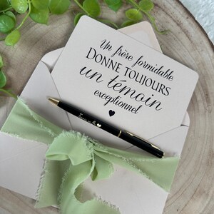 May include: A light pink envelope with a light green ribbon and a black pen. A card inside the envelope has text in French. The pen has gold accents and a small heart. Green leaves are in the background.