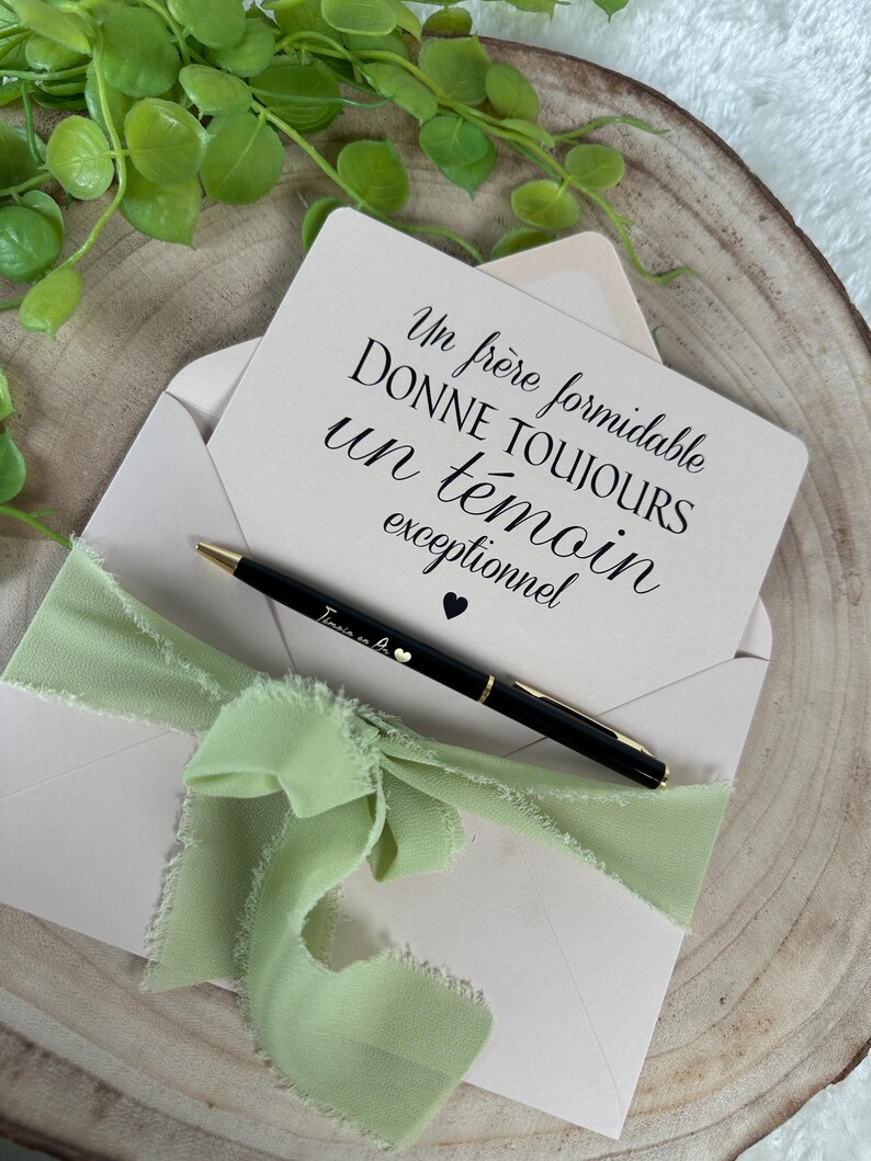 May include: A light pink envelope with a light green ribbon and a black pen. A card inside the envelope has text in French. The pen has gold accents and a small heart. Green leaves are in the background.