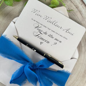 May include: A cream-colored card inside a white envelope, tied with a bright blue ribbon. The card has elegant script that reads "Mon Meilleur Ami, Je ne peux pas dire "oui" sans toi &agrave; mes c&ocirc;t&eacute;s, Veux-tu &ecirc;tre mon T&eacute;moin ?" A black pen with gold accents rests on the envelope.