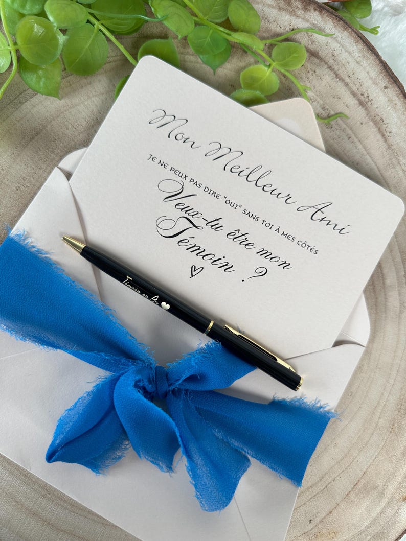 May include: A cream-colored card inside a white envelope, tied with a bright blue ribbon. The card has elegant script that reads "Mon Meilleur Ami, Je ne peux pas dire "oui" sans toi &agrave; mes c&ocirc;t&eacute;s, Veux-tu &ecirc;tre mon T&eacute;moin ?" A black pen with gold accents rests on the envelope.