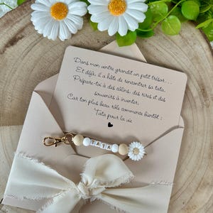 Puede incluir: Una tarjeta beige con texto en franc&eacute;s, un llavero dorado con la palabra "TATA" escrita con cuentas blancas y de madera, y un dije de margarita blanca. La tarjeta est&aacute; dentro de un sobre beige con una cinta color crema.