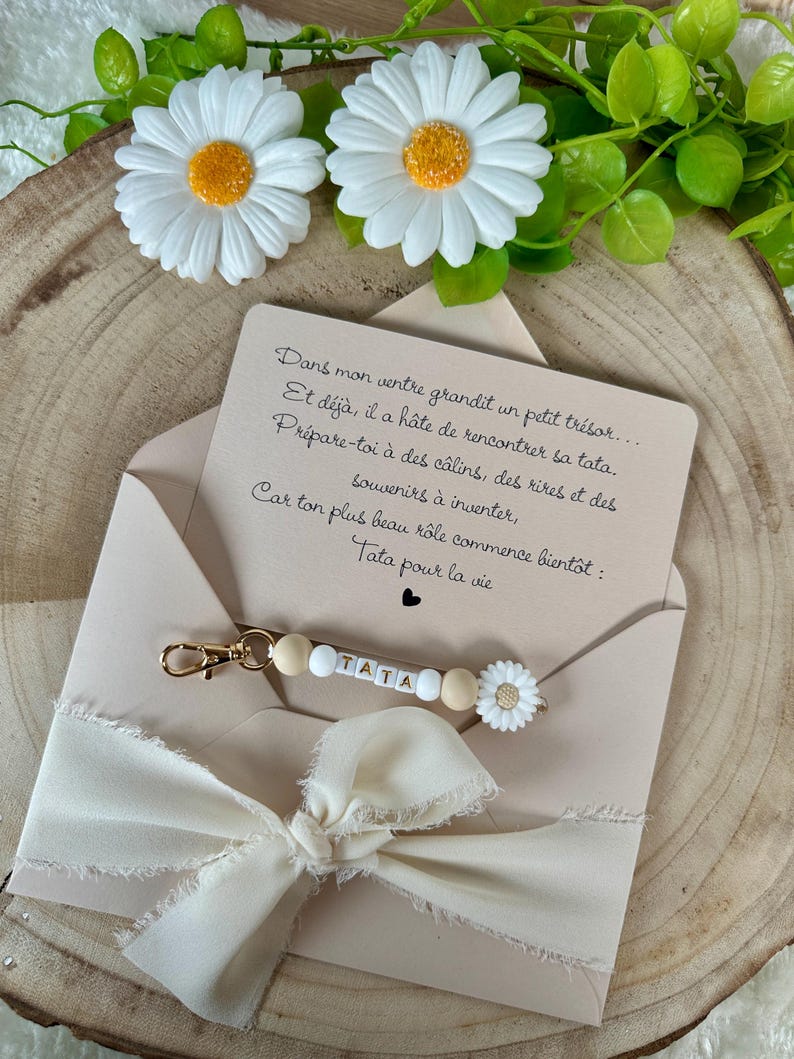Puede incluir: Una tarjeta beige con texto en franc&eacute;s, un llavero dorado con la palabra "TATA" escrita con cuentas blancas y de madera, y un dije de margarita blanca. La tarjeta est&aacute; dentro de un sobre beige con una cinta color crema.