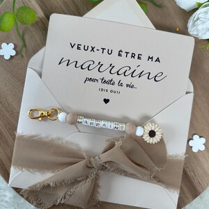 以下が含まれることがあります： 白いカードに黒い文字で「VEUX-TU &Ecirc;TRE MA marraine pour toute la vie... (DIS OUI)」と書かれています。カードの周りに、ほつれた縁のある茶色のリボンが結ばれています。リボンの上に、金色の文字で「MARRAINE」と綴られた白と茶色のビーズキーホルダーがあります。