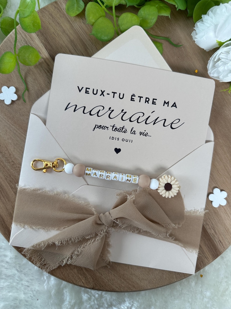 以下が含まれることがあります： 白いカードに黒い文字で「VEUX-TU &Ecirc;TRE MA marraine pour toute la vie... (DIS OUI)」と書かれています。カードの周りに、ほつれた縁のある茶色のリボンが結ばれています。リボンの上に、金色の文字で「MARRAINE」と綴られた白と茶色のビーズキーホルダーがあります。