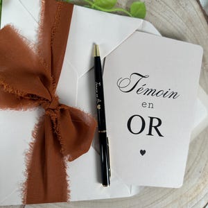 May include: A white envelope tied with a rust-colored ribbon, a black pen, and a card with the words "T&eacute;moin en OR" printed on it. The pen has gold accents and the text "T&eacute;moin en Or" printed on it.