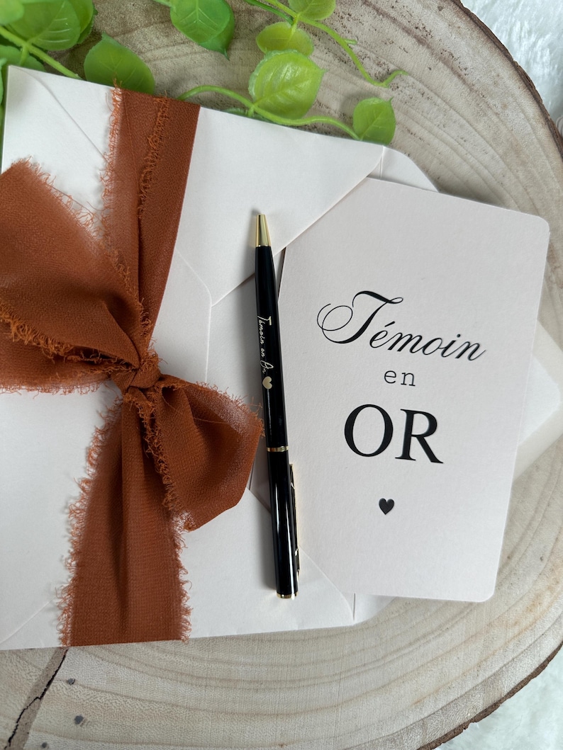 May include: A white envelope tied with a rust-colored ribbon, a black pen, and a card with the words "T&eacute;moin en OR" printed on it. The pen has gold accents and the text "T&eacute;moin en Or" printed on it.