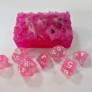 Könnte beinhalten: Ein Set aus rosa Kunstharz-Würfeln mit weißen Zahlen und Glitzerakzenten. Die Würfel sind um eine passende rosa Kunstharzbox mit Rosenmuster angeordnet. Das Set enthält verschiedene Formen für Tischspiele.