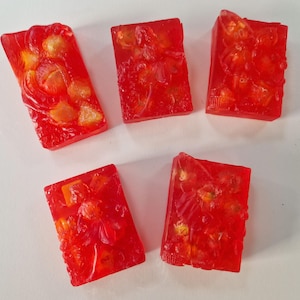 Könnte beinhalten: Fünf rote, rechteckige, durchscheinende Seifenstücke mit einem Blumendesign. Die Seifenstücke haben eine strukturierte Oberfläche und sind mit kleinen, gelben Flocken gefüllt.