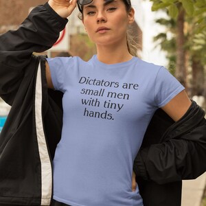以下が含まれることがあります： 黒い文字で「Dictators are small men with tiny hands.」と書かれた薄い青色のTシャツです。