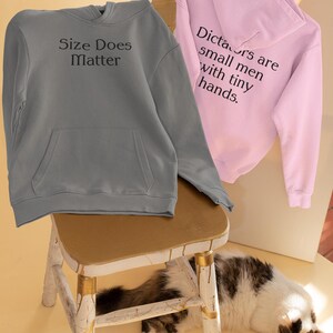 以下が含まれることがあります： フード付きスウェットシャツ2枚。グレーのものは「Size Does Matter」と書いてあり、ピンクのものは「Dictators are small men with tiny hands.」と書いてある。