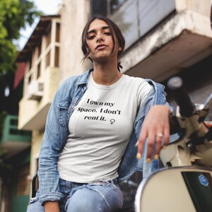 Puede incluir: Una mujer con una camiseta blanca con el texto "I own my space. I don't rent it. ♀" impreso en ella. Lleva una chaqueta vaquera y unos vaqueros azules.