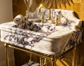 May include: A white and purple marble sink basin with a gold metal faucet and base. The sink features a matching marble shelf below, a rolled beige towel, and a small marble dish. The wall behind is covered in square mustard yellow tiles.