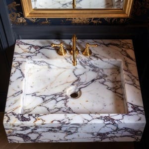 May include: A rectangular white marble sink featuring prominent dark purple and grey veining throughout the stone. The basin is equipped with a polished gold-toned faucet and two matching gold-toned handles mounted on the flat rear deck.