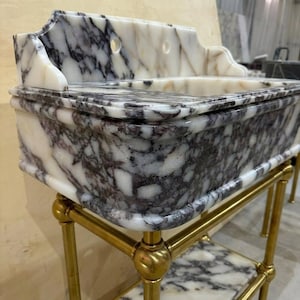 May include: A rectangular marble sink featuring a white base with dark purple and grey veining. The sink has a high back splash with two circular faucet holes and rests on a gold-toned metal stand with a matching marble shelf at the bottom.