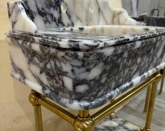 May include: A rectangular marble sink featuring a white base with dark purple and grey veining. The sink has a high back splash with two circular faucet holes and rests on a gold-toned metal stand with a matching marble shelf at the bottom.
