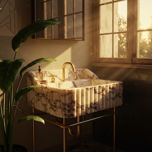 May include: A marble bathroom sink with gold fixtures. The sink has a rectangular basin with a fluted design and is supported by a gold metal frame. A soap dispenser sits on the sink's edge, and a large plant is in the foreground.