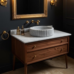 May include: A bathroom vanity featuring a marble countertop and a grey, ribbed vessel sink. The vanity is crafted from dark wood, with two drawers and brass hardware. A gold-framed mirror hangs above, complemented by gold fixtures and accessories.