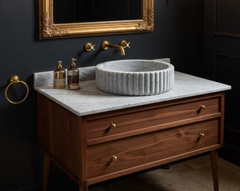 May include: A bathroom vanity featuring a dark wood cabinet with two drawers, a white marble countertop, and a fluted circular marble vessel sink. The setup includes a gold faucet, two gold wall sconces, a gold-framed mirror, and a gold towel ring.