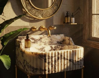 May include: A marble bathroom sink with a gold faucet and fixtures. The sink has a patterned design and is supported by gold legs. A gold-framed mirror hangs above the sink, and a bottle of soap sits on the counter.