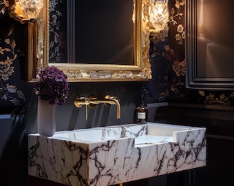 Può includere: Un lavabo da bagno in marmo bianco con un rubinetto dorato e uno specchio con cornice dorata con una carta da parati floreale nera e dorata dietro.
