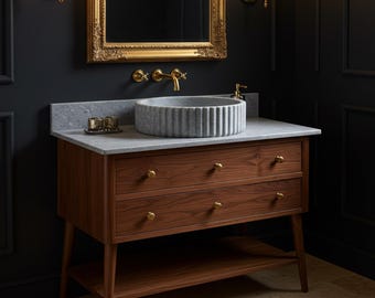 May include: A wooden bathroom vanity with a gray marble countertop and a round, ribbed stone sink. The vanity has two drawers and a lower shelf, with brass hardware and legs. A gold-framed mirror and sconces are mounted on the dark wall.