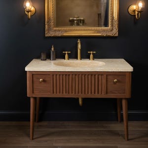 May include: A wooden bathroom vanity with a light-colored stone countertop and a built-in oval sink. The vanity has two drawers with brass knobs and a ribbed center panel. A gold-colored faucet and mirror are above the vanity.